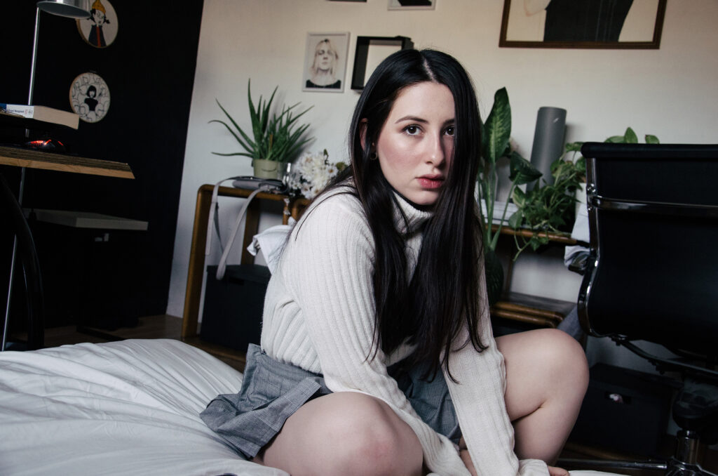 Gabriela Inzunza sits on a bed in a room adorned with artwork plants and a desk with a computer.